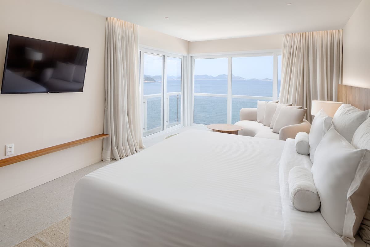 A large bed with white linens, a flat-screen TV mounted on the wall, and a view of the sea through large windows.