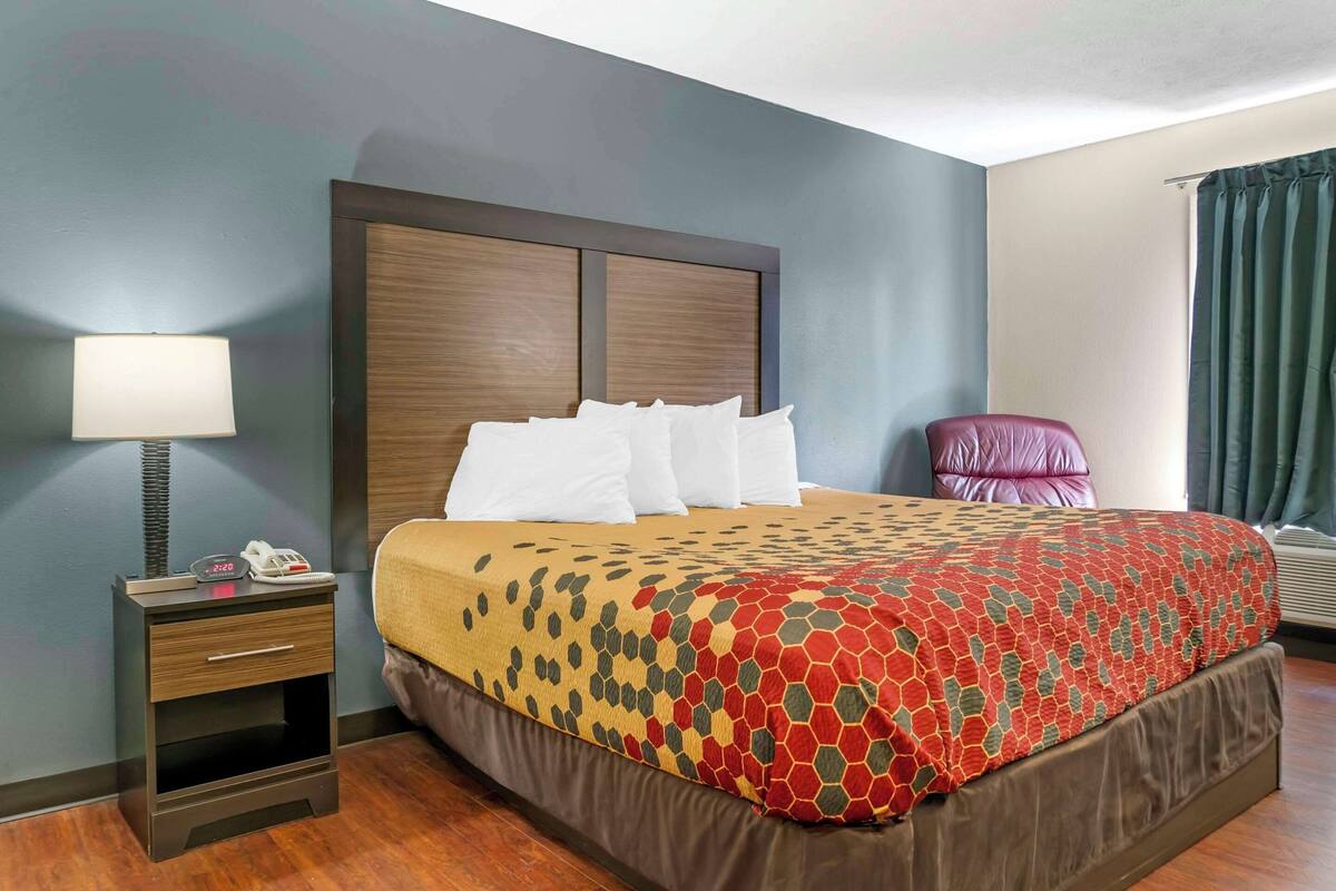 A hotel room with a large bed, a bedside table with a lamp, a chair, and a window with curtains.