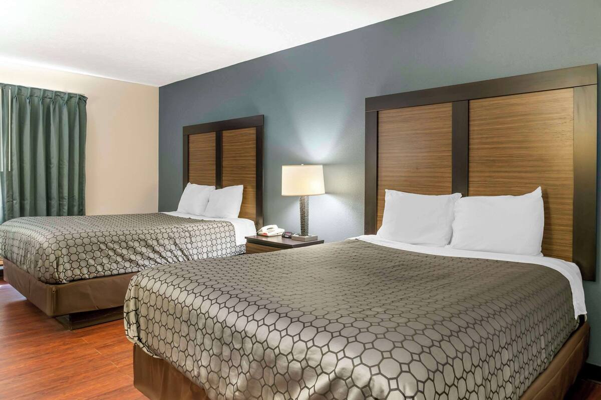 A hotel room with two beds, each with a patterned bedspread, a bedside table with a lamp, and a curtain on one side.