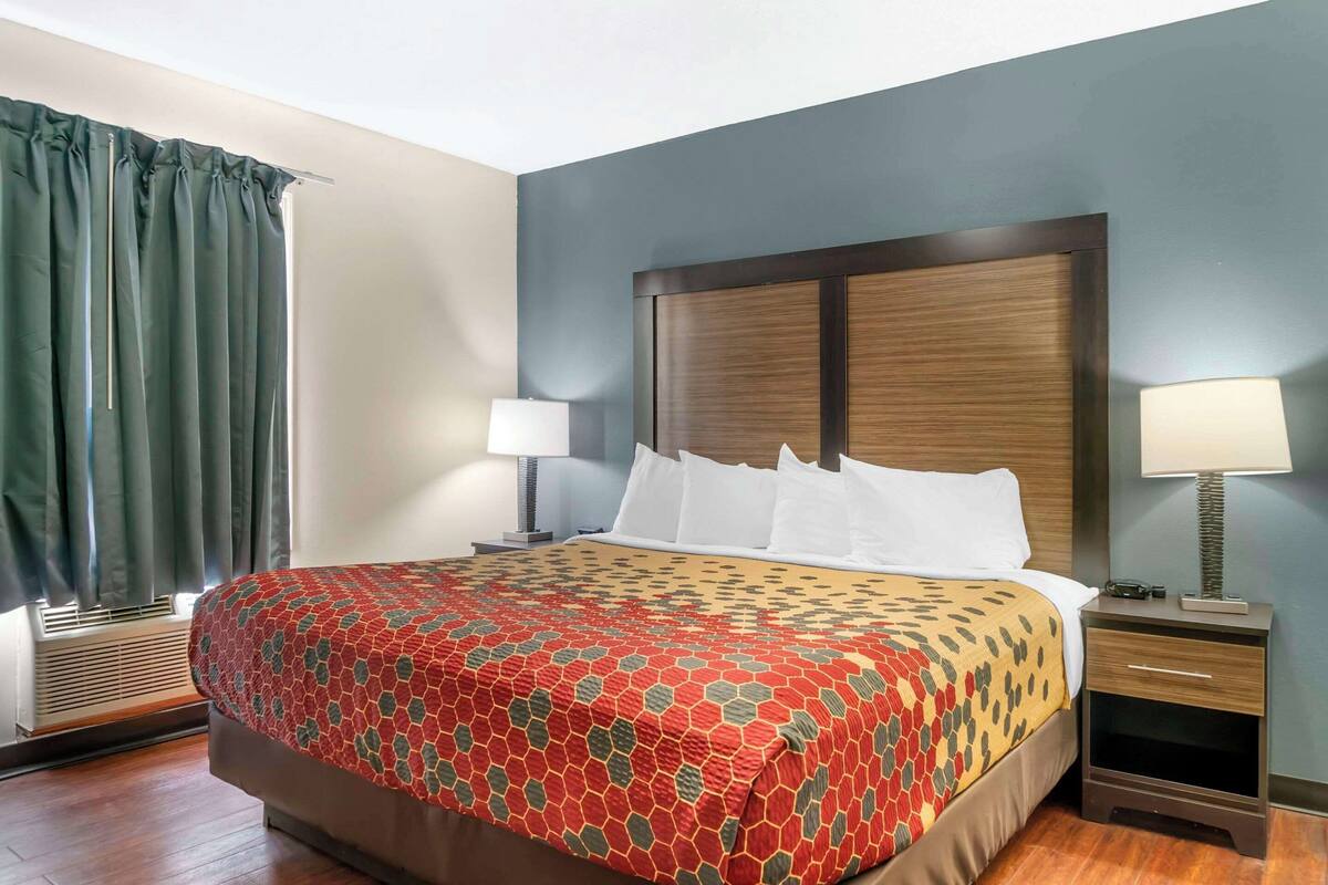 A hotel room with a large bed, two bedside tables with lamps, a window with curtains, and a wall-mounted air conditioning unit.