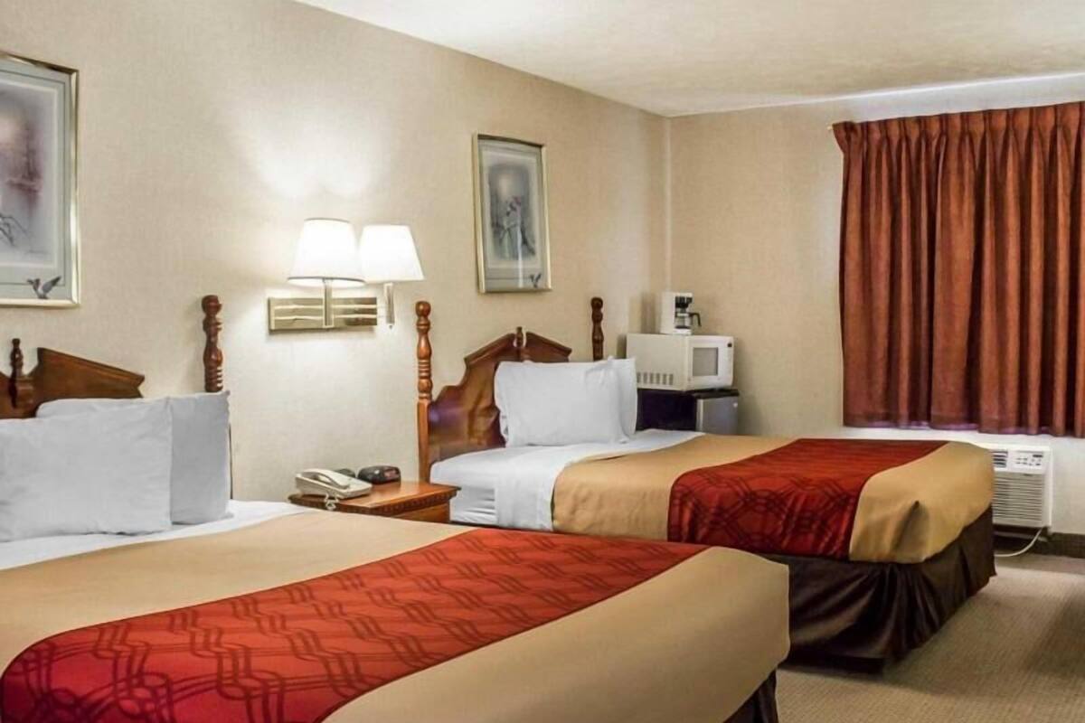 A hotel room with two beds, a bedside table with a lamp, a microwave, and a window with curtains.