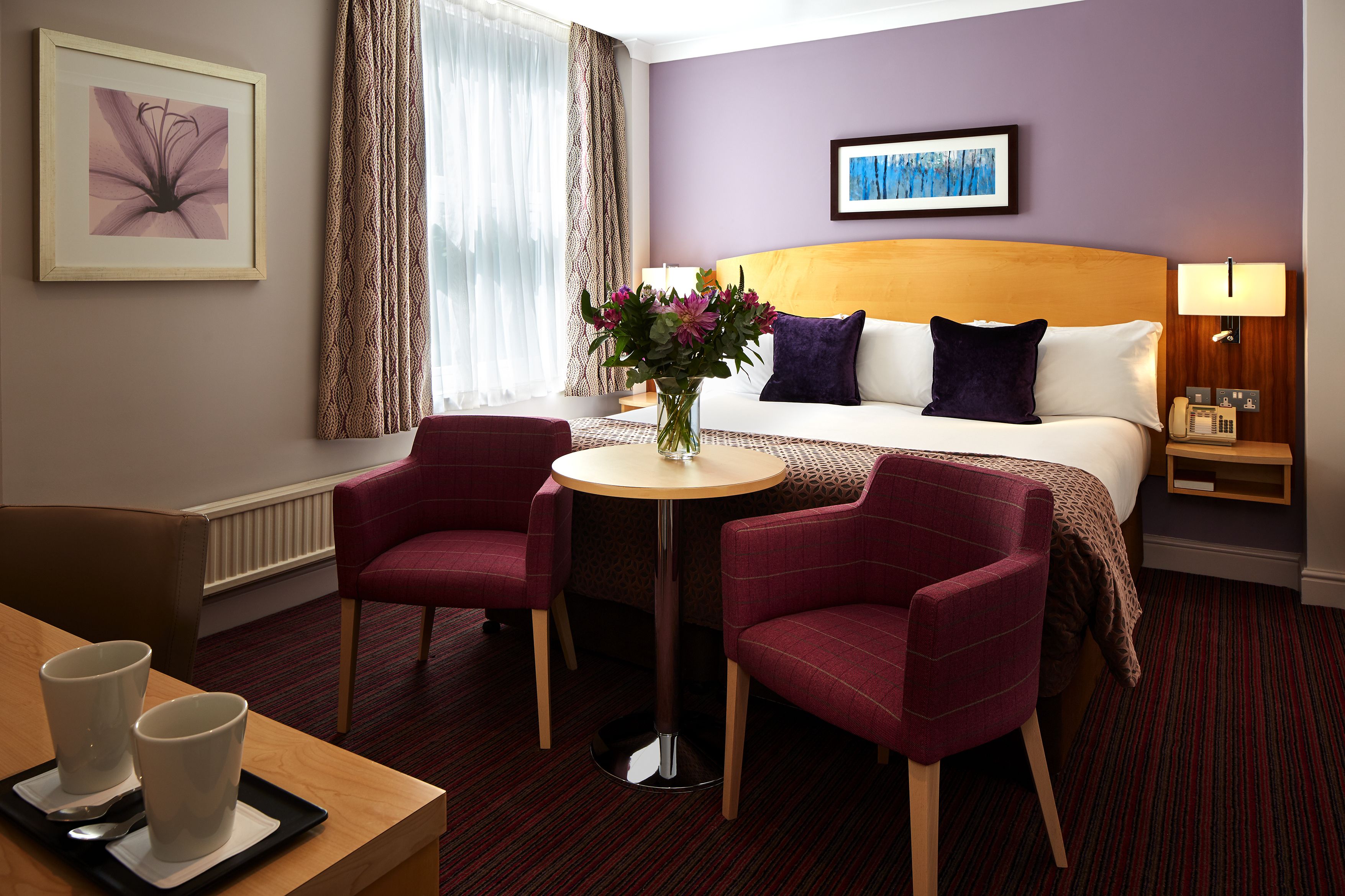 A hotel room with a bed, two chairs, a small table with cups, a vase with flowers, and artwork on the walls.