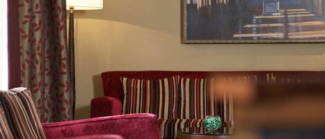 A hotel lobby with a red sofa, striped armchairs, a lamp, and a painting of a library scene.