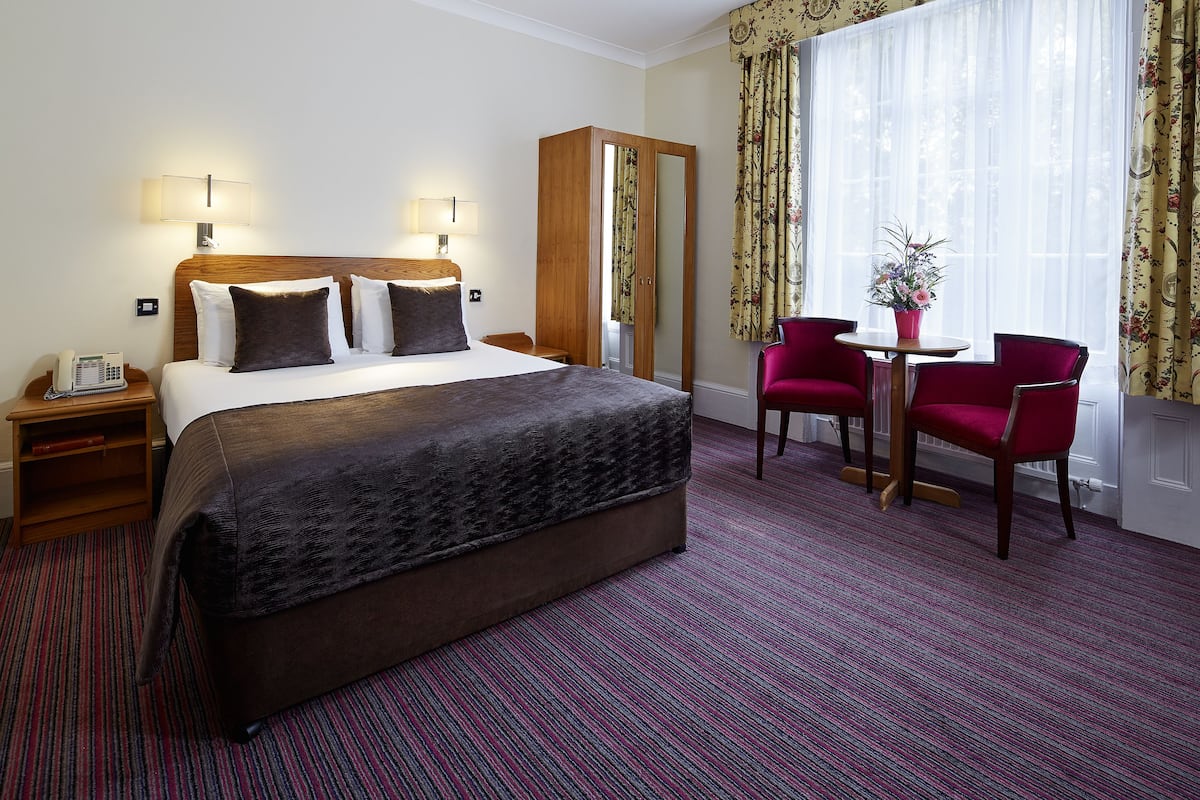Quarto de hotel com uma cama grande, luminárias de cabeceira, um guarda-roupa, uma escrivaninha com telefone, uma mesinha com cadeira e uma janela com cortinas.
