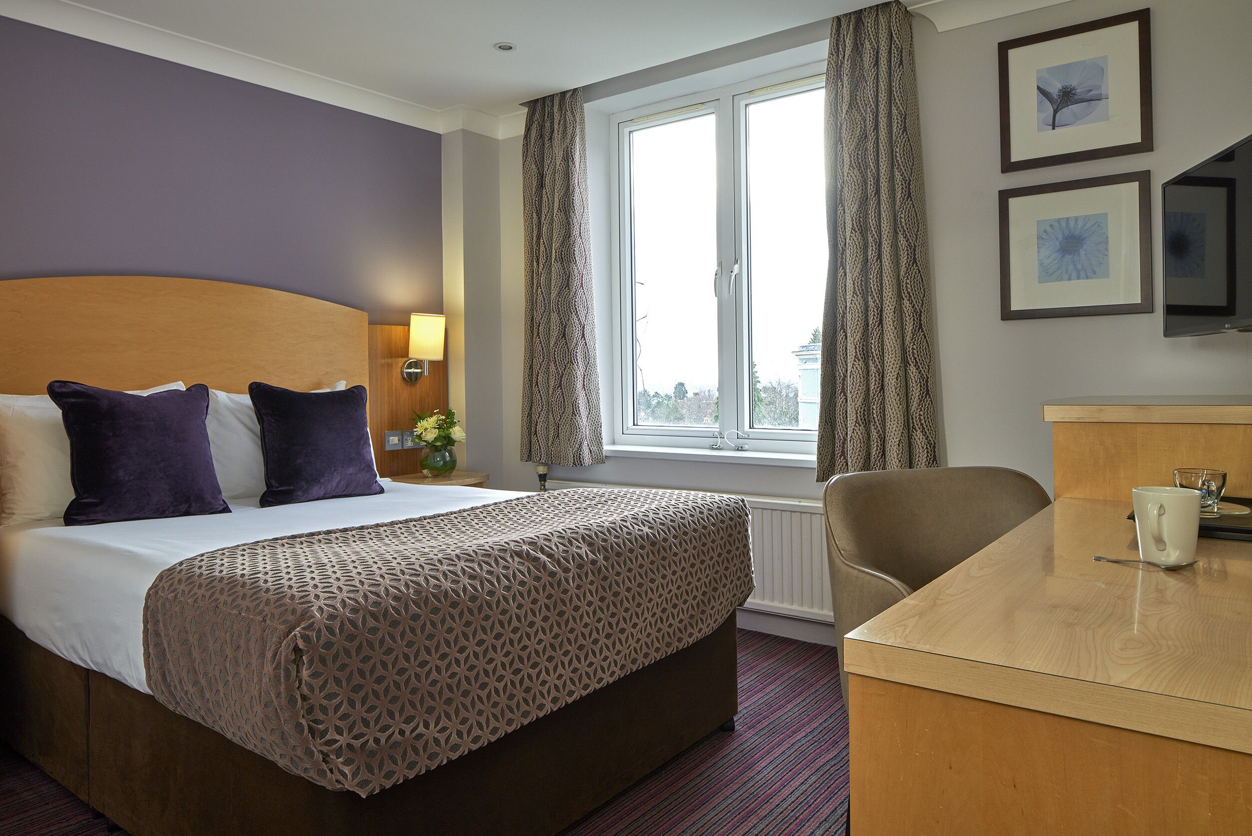 A hotel room with a bed, a desk, a chair, a window with curtains, and framed pictures on the wall.