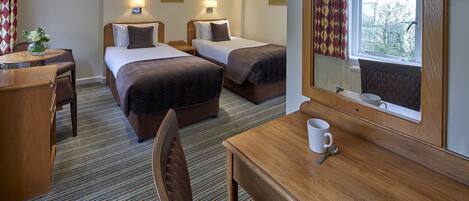 A hotel room with two beds, a wooden desk, a chair, a table with a cup, and a window with curtains.