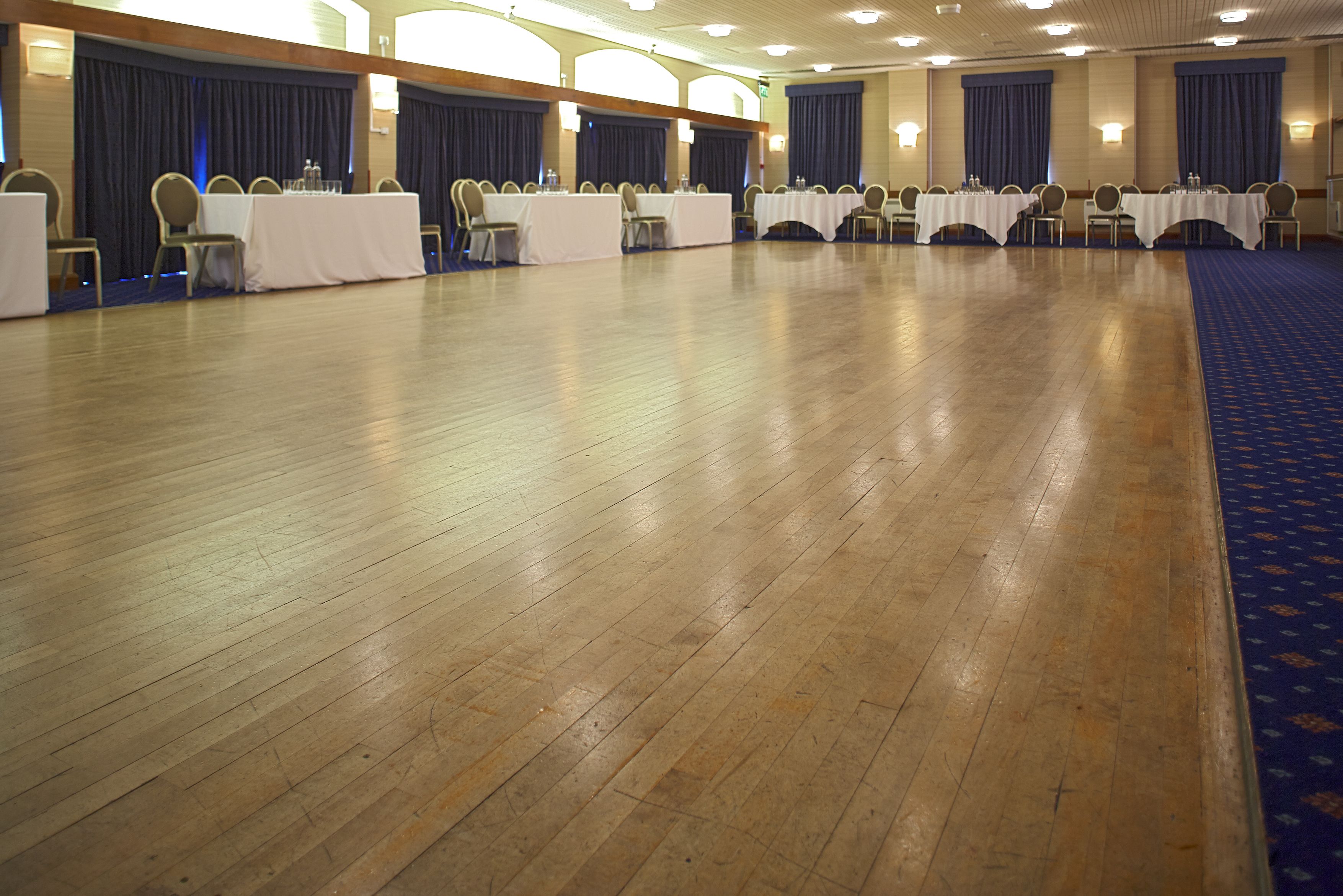 A conference room with multiple tables and chairs, wooden floors, and blue curtains.