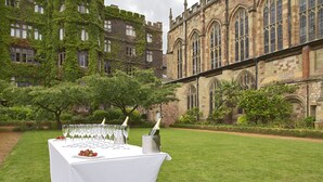 A table set outdoors with champagne glasses and a bottle, in front of a historic building with ivy-covered walls. - The Abbey Great Malvern (Malvern)