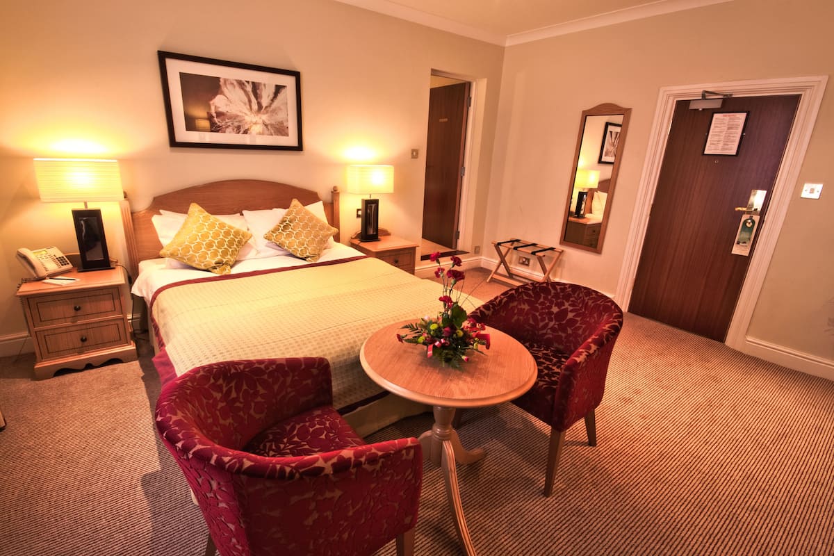 Quarto de hotel com uma cama, duas mesas de cabeceira, uma mesinha de apoio, uma mesa redonda de madeira com cadeiras e um espelho na parede.