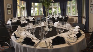 A round dining table set for a formal event with multiple chairs, a central floral arrangement, and a view of the outdoors through large windows. - The Abbey Great Malvern (Malvern)