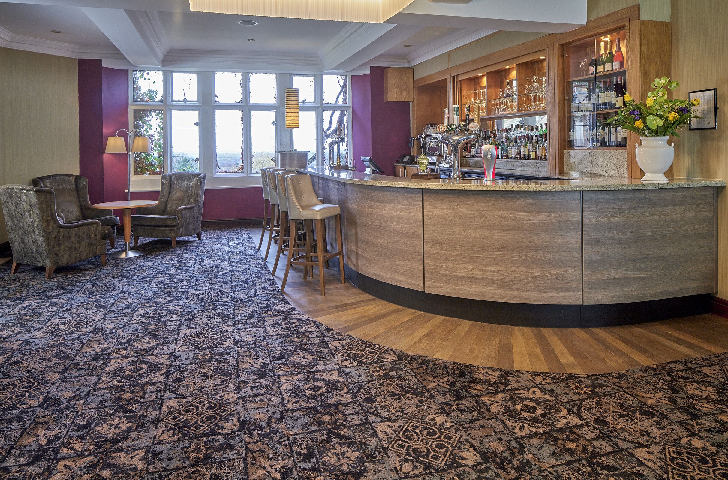A bar area with a curved counter, bar stools, and a variety of bottles and glasses. There are chairs and a patterned carpet.
