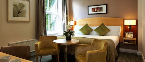 A hotel room with a bed, two chairs, a small table, a window with curtains, and a framed picture on the wall.