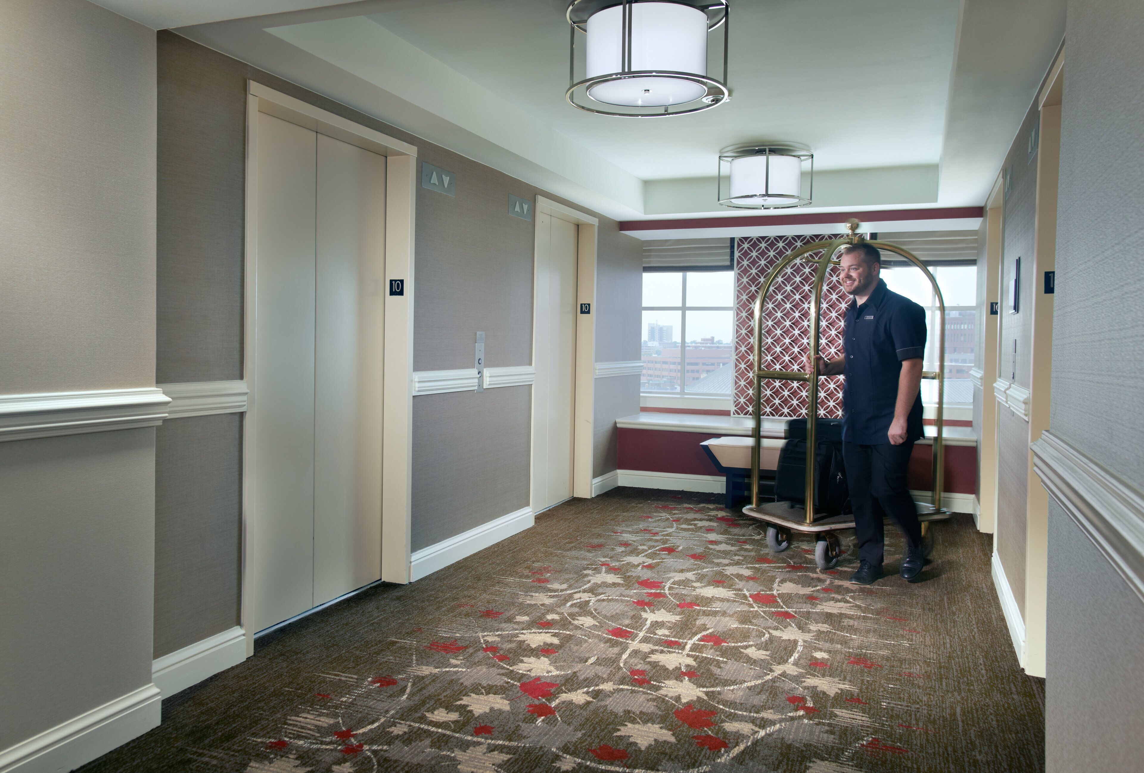 A hotel hallway with a carpeted floor, a luggage cart, and a window with a view of buildings.
