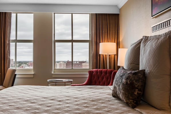 A hotel room with a large bed, a red armchair, a small table, and a view of the city through large windows.