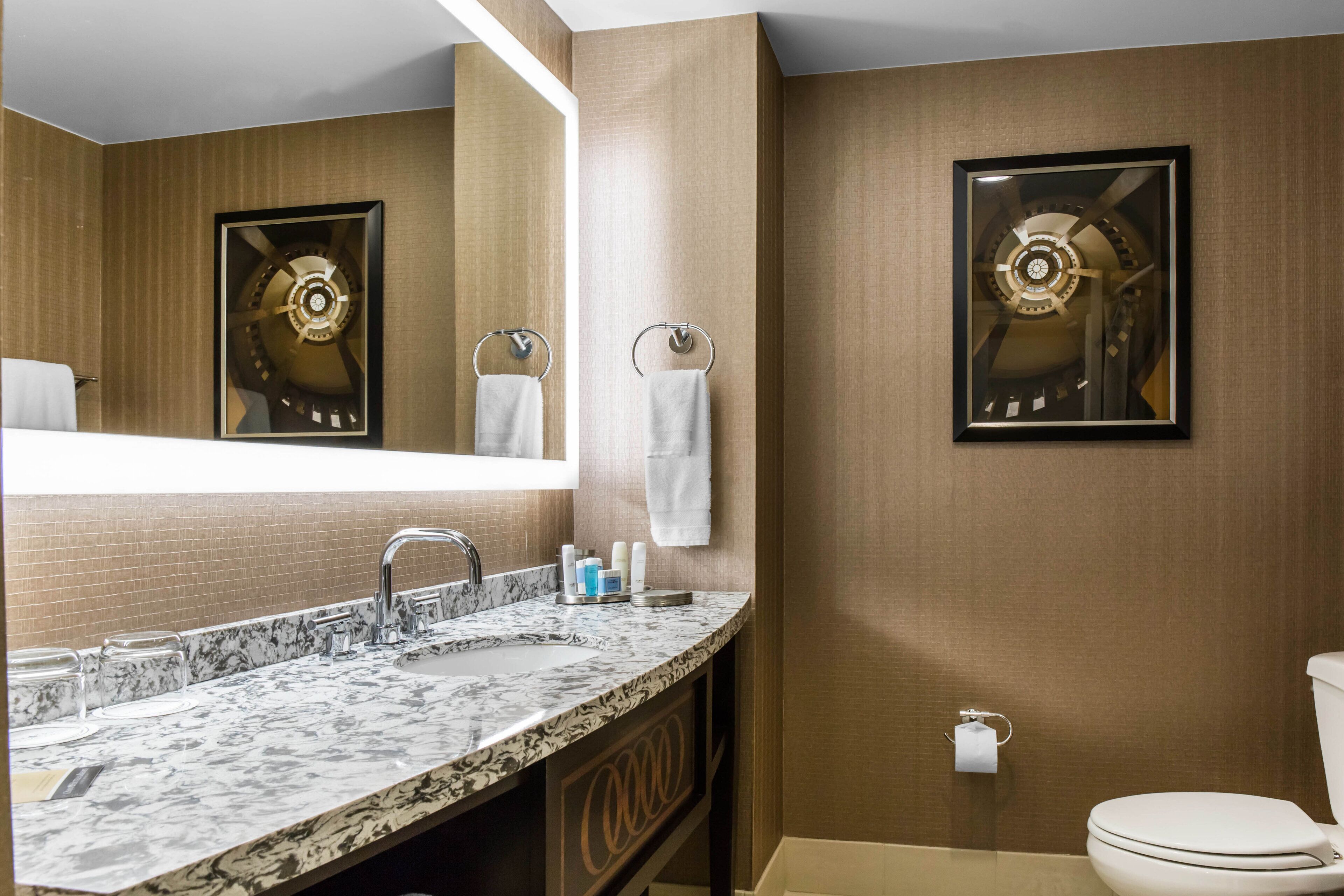 A modern bathroom with a marble sink, a large mirror, a toilet, and a framed clock on the wall.