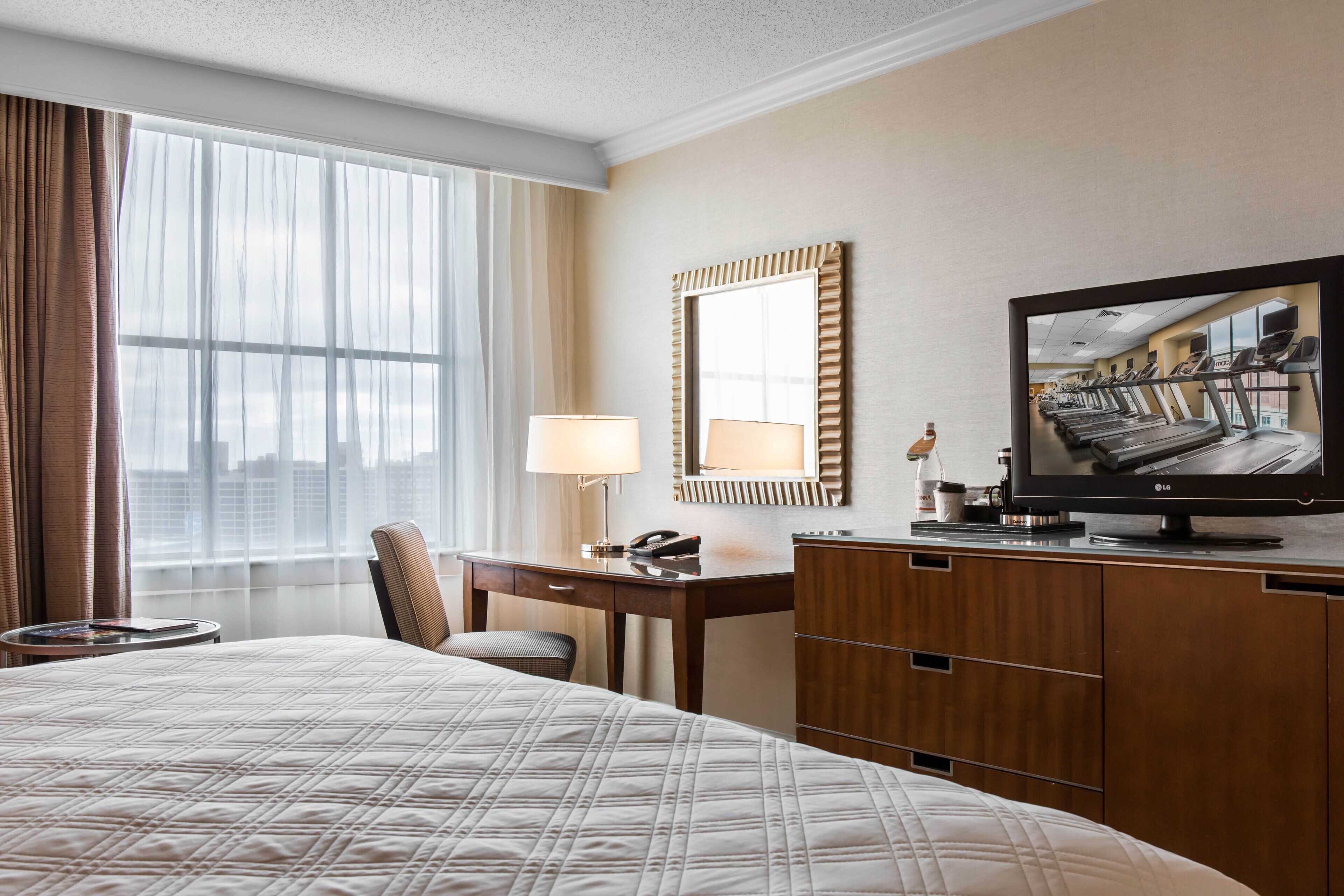 A hotel room with a large bed, a desk with a lamp, a television, and a view of the city through large windows.