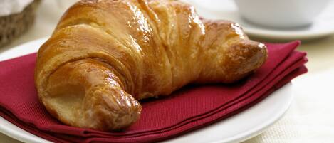 A croissant on a plate with a red napkin, a glass of orange juice, a cup of coffee, and a basket with an apple and a tomato in the background.