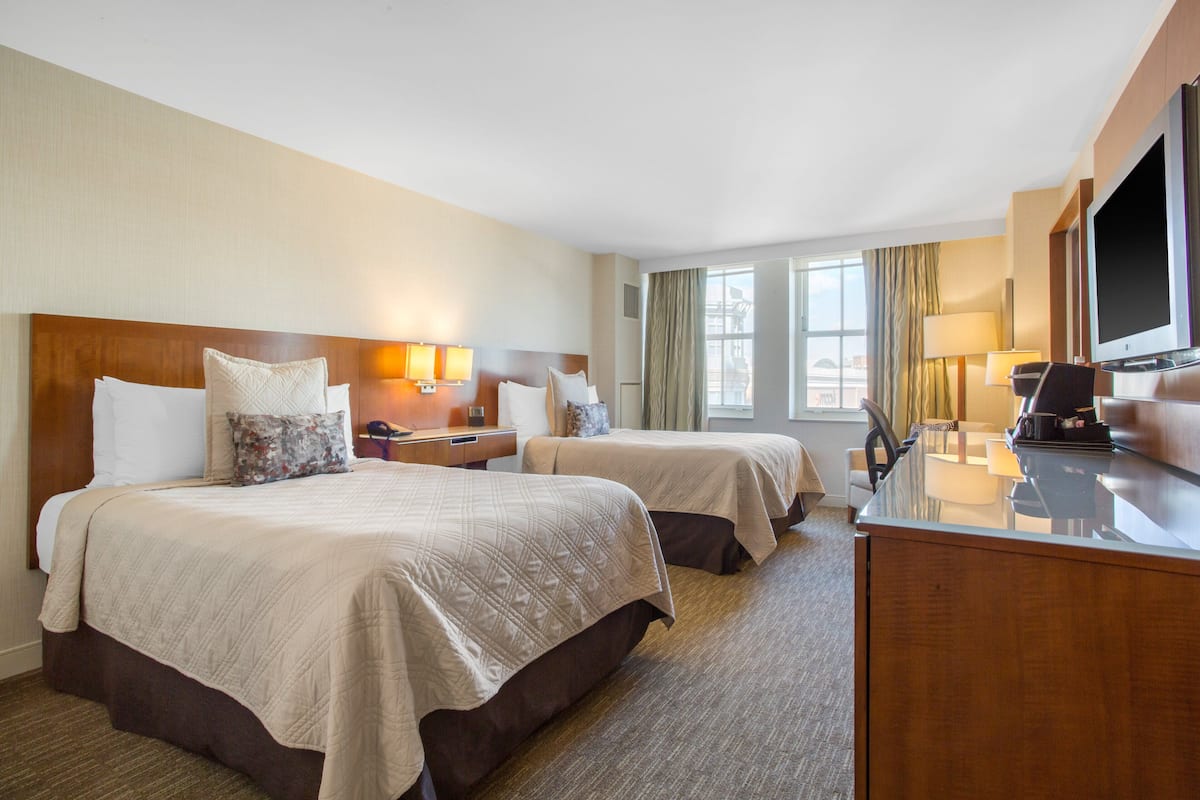 A hotel room with two beds, a desk, a television, and a window with curtains.