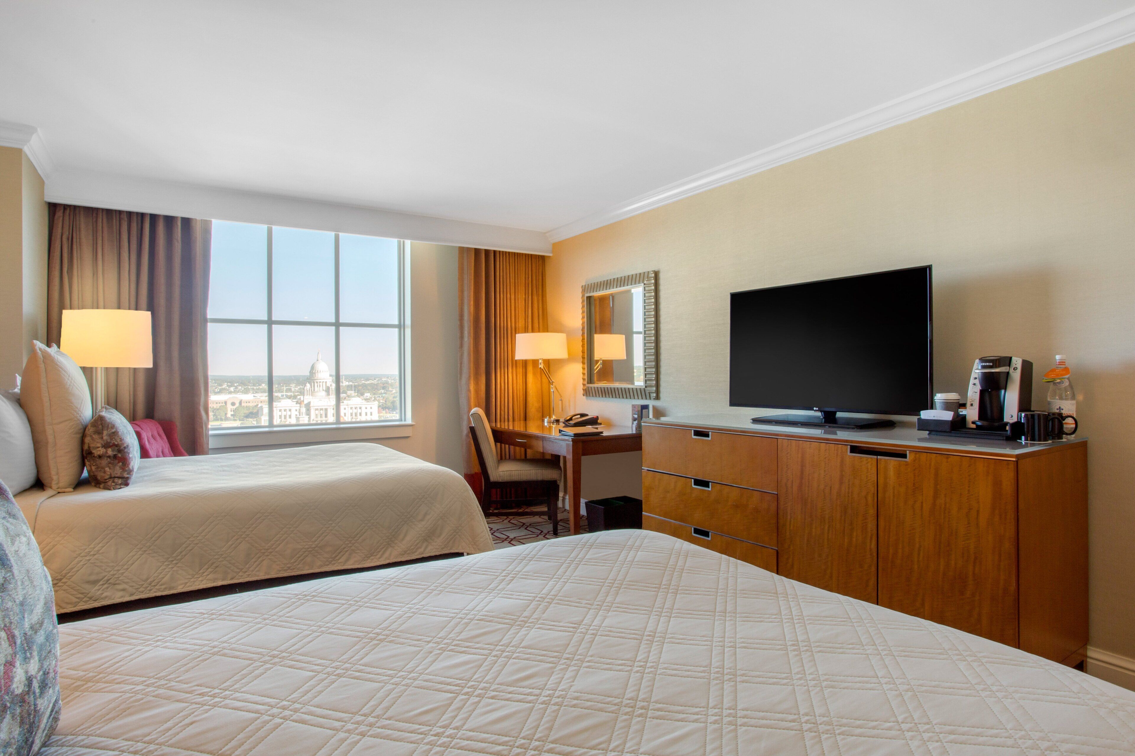 A hotel room with two beds, a desk, a television, and a view of the city.