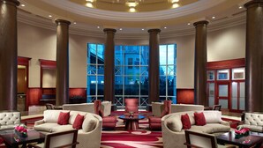 Un vestíbulo espacioso con techos altos, amplias columnas y un gran candelabro. Hay varias zonas de estar con sofás y sillones, y grandes ventanales que ofrecen vistas de la ciudad.