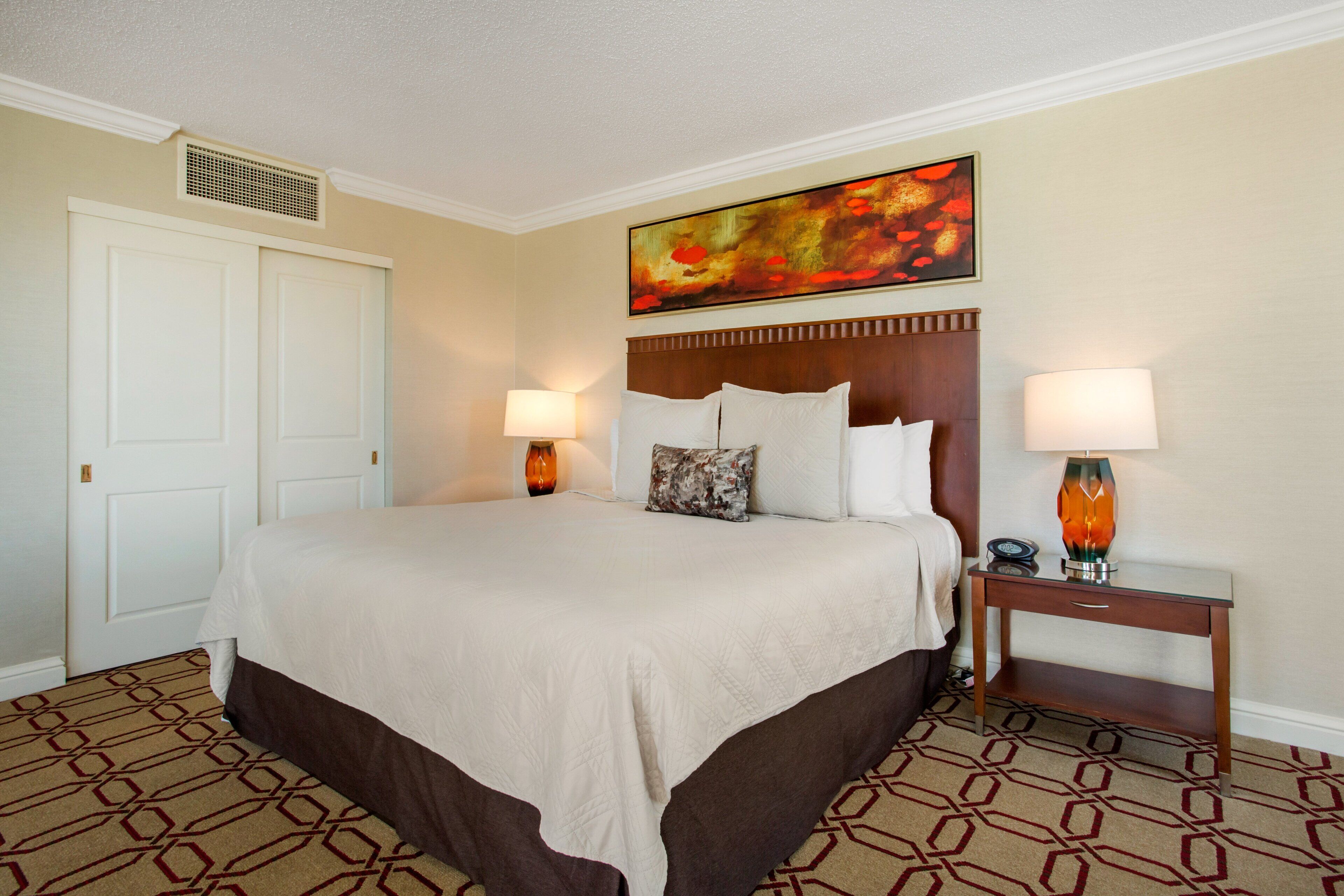 A hotel room with a large bed, two bedside lamps, a headboard, a painting above the bed, and a patterned carpet.