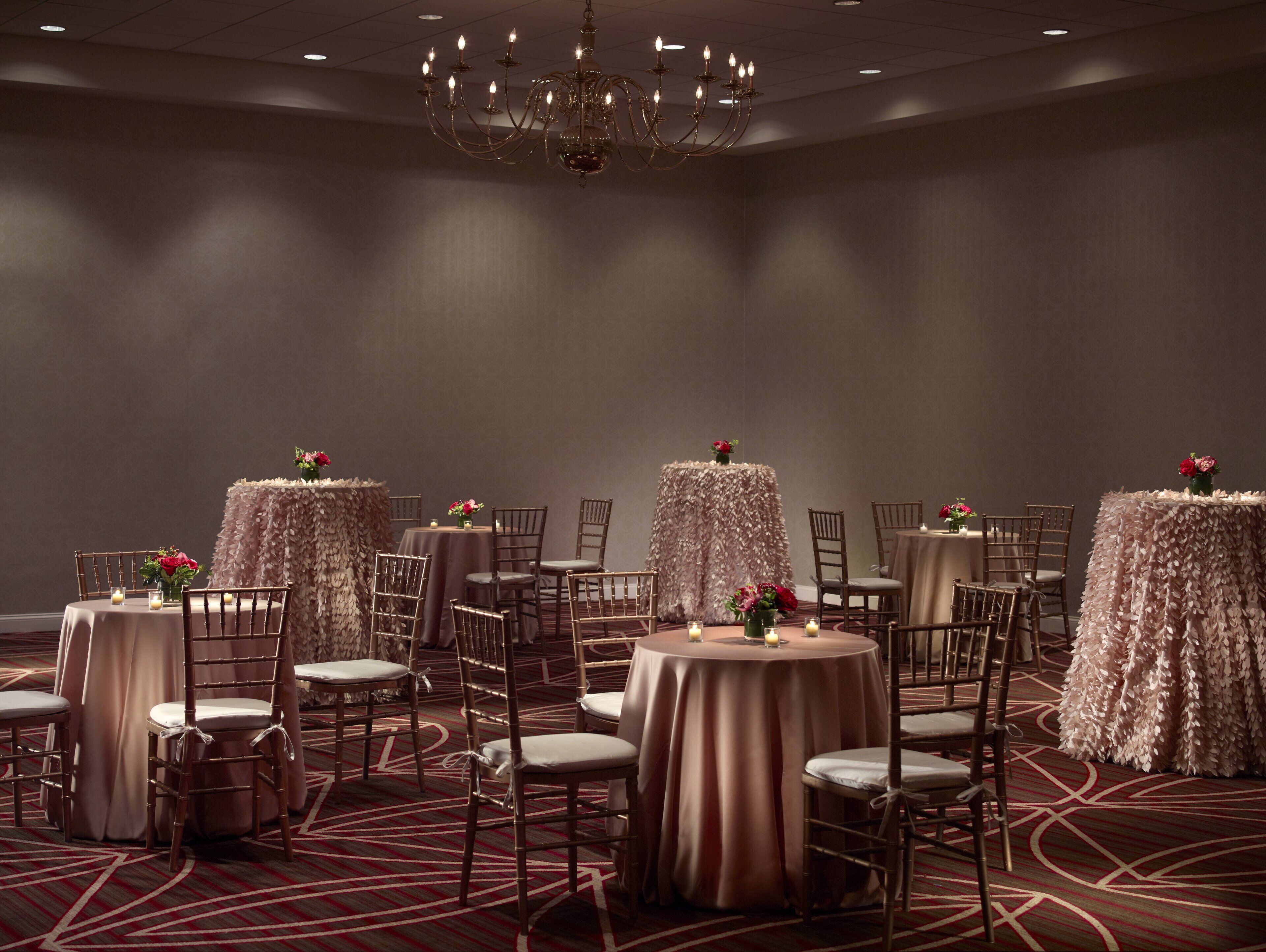 A banquet hall with round tables covered in tablecloths, surrounded by chairs, and a chandelier hanging from the ceiling.