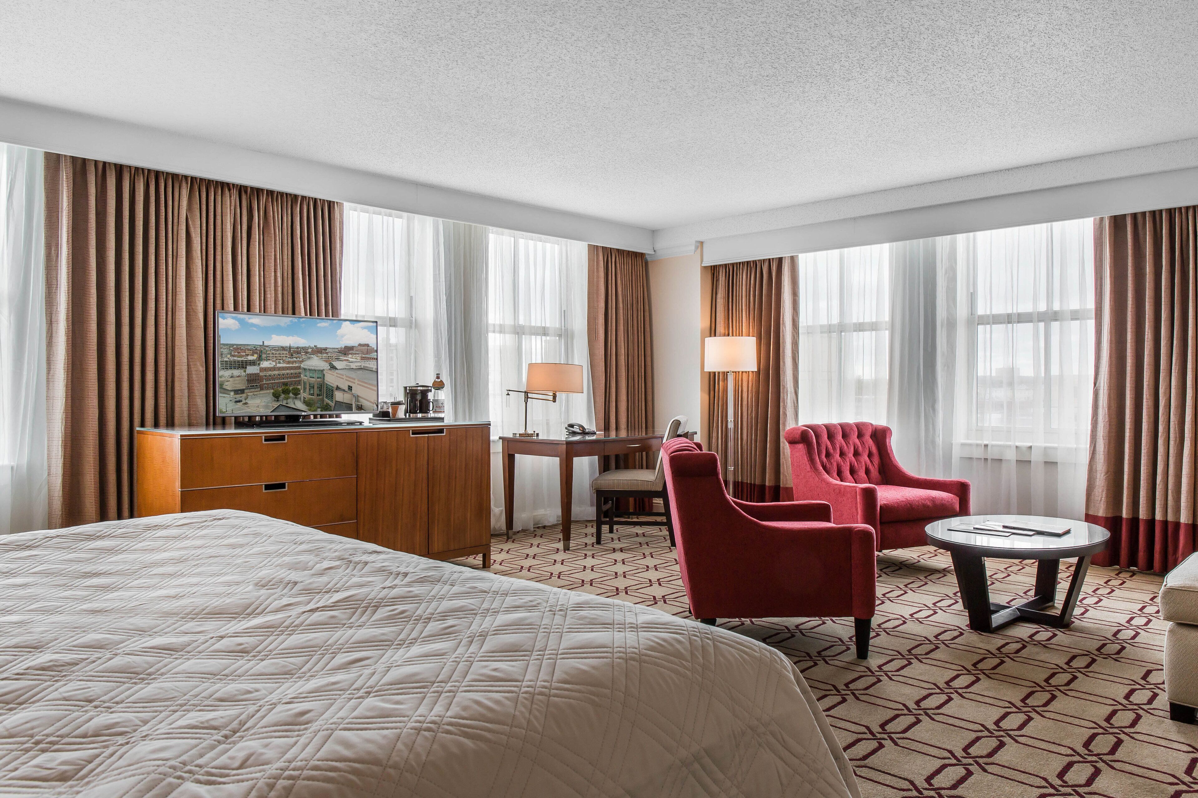 A hotel room with a large bed, a desk with a chair, a red armchair, a small round table, and a television on a dresser.