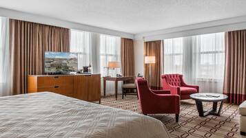 A hotel room with a large bed, a desk with a chair, a red armchair, a small round table, and a television on a dresser.