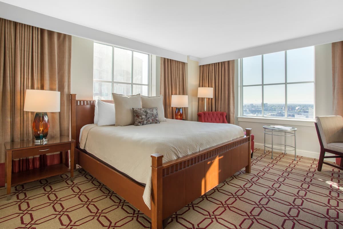 A hotel room with a large bed, two bedside tables with lamps, a small side table, a chair, and a view of the city through large windows.