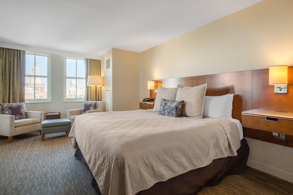 A hotel room with a large bed, two bedside tables with lamps, a chair, and a view of a building through the window.