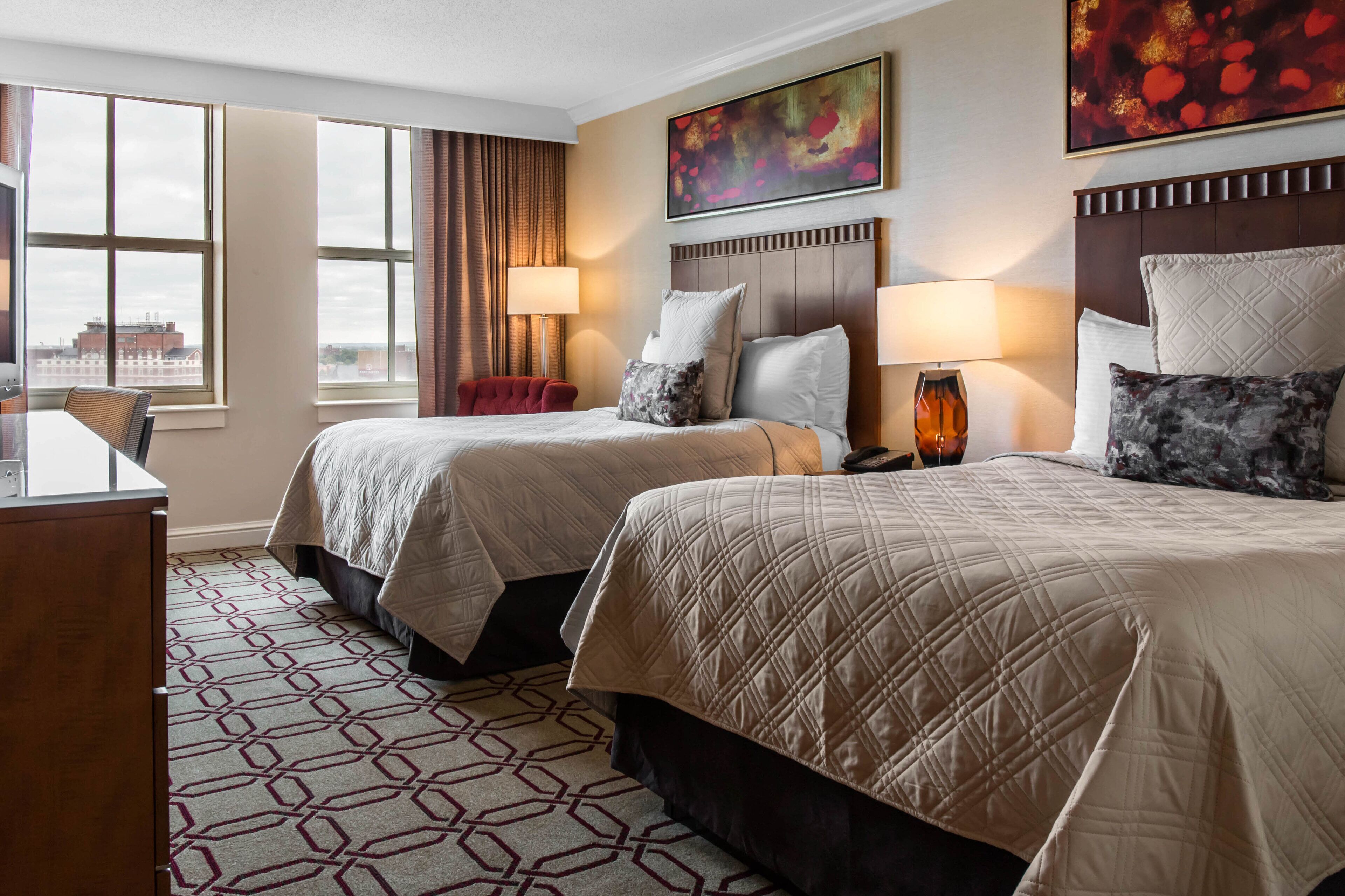 A hotel room with two beds, a desk, and a view of the city.