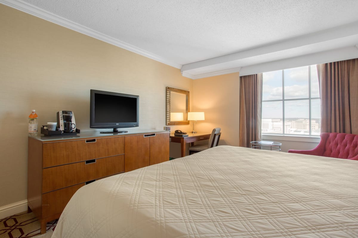 A hotel room with a bed, a desk, a television, a lamp, a chair, and a view of the city through the window.