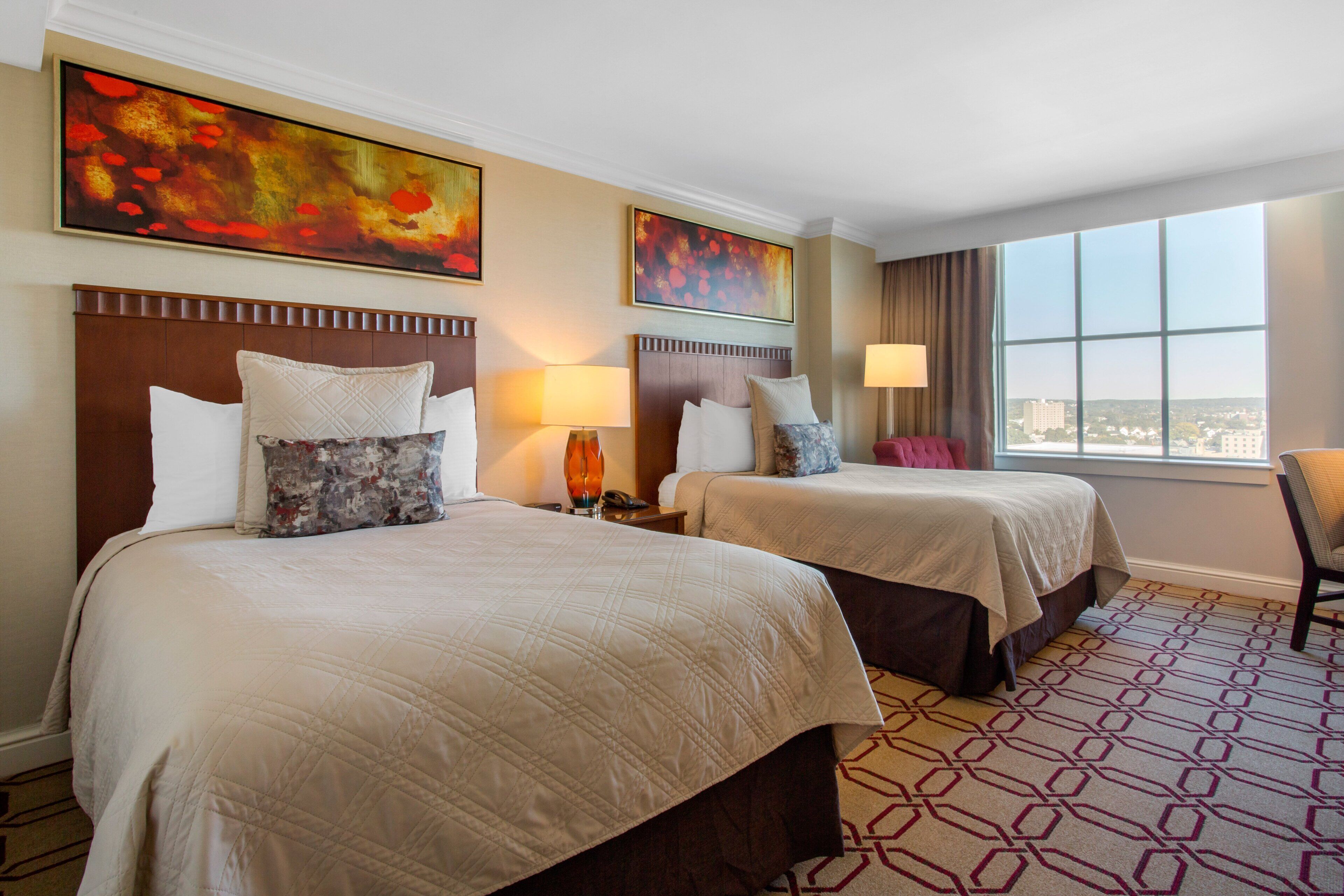 A hotel room with two beds, a large window, and artwork on the walls.