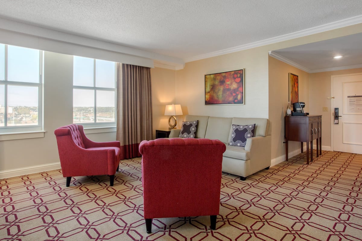 A hotel room with a large window, a sofa, two armchairs, a lamp, and a painting on the wall.