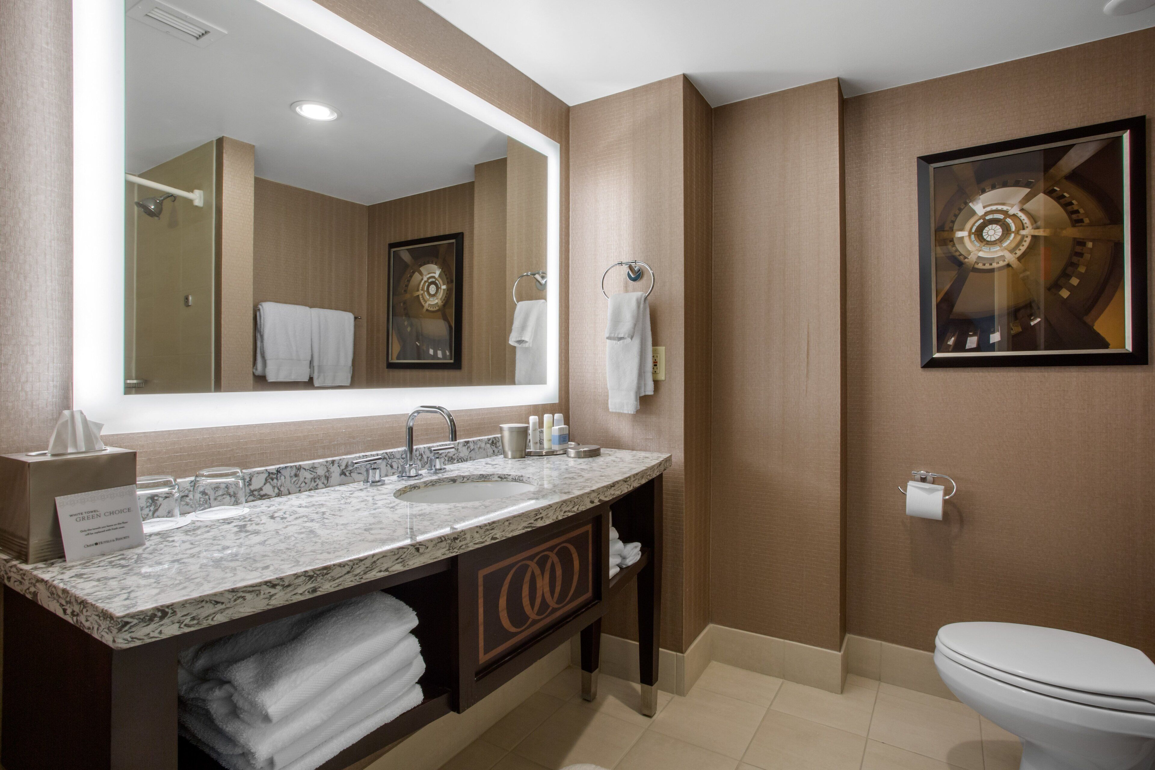 A modern bathroom with a large mirror, dual sinks, a toilet, and a framed artwork on the wall.