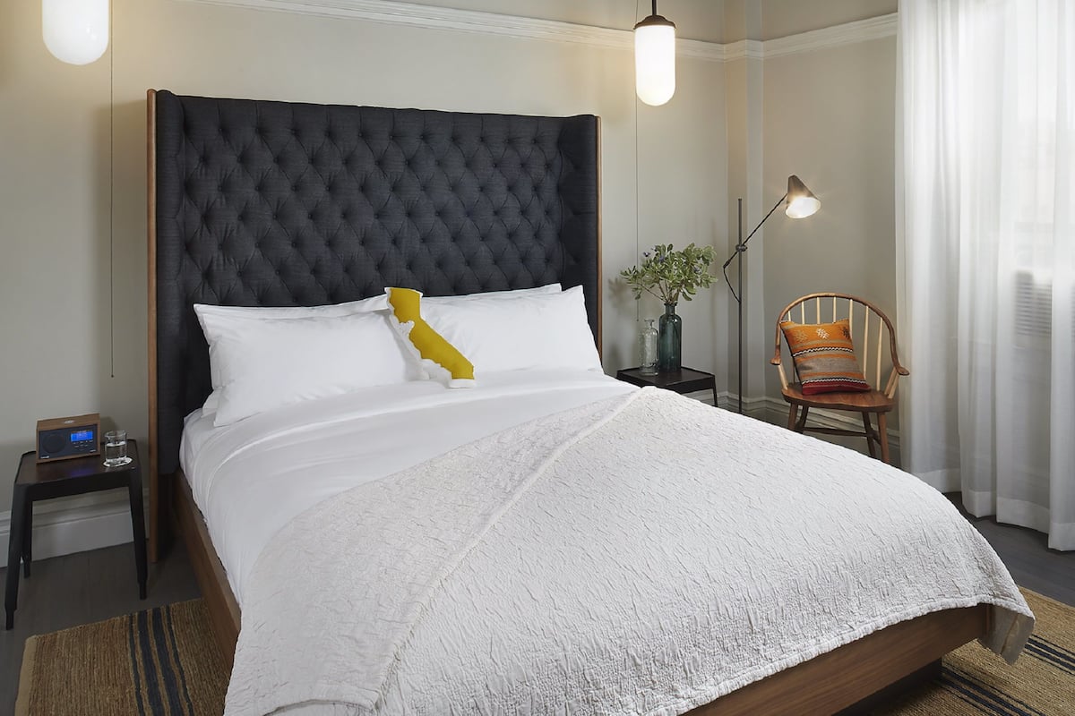 A neatly made bed with a tufted headboard, white bedding, and a single decorative pillow. There is a bedside table with a radio and a glass of water. A floor lamp and a chair with a cushion are also visible.