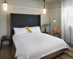 Una cama bien tendida con cabecera capitonada, ropa de cama blanca y una única almohada decorativa. Se observa una mesita de noche con una radio y un vaso de agua. También se ve una lámpara de pie y un sillón con cojín.