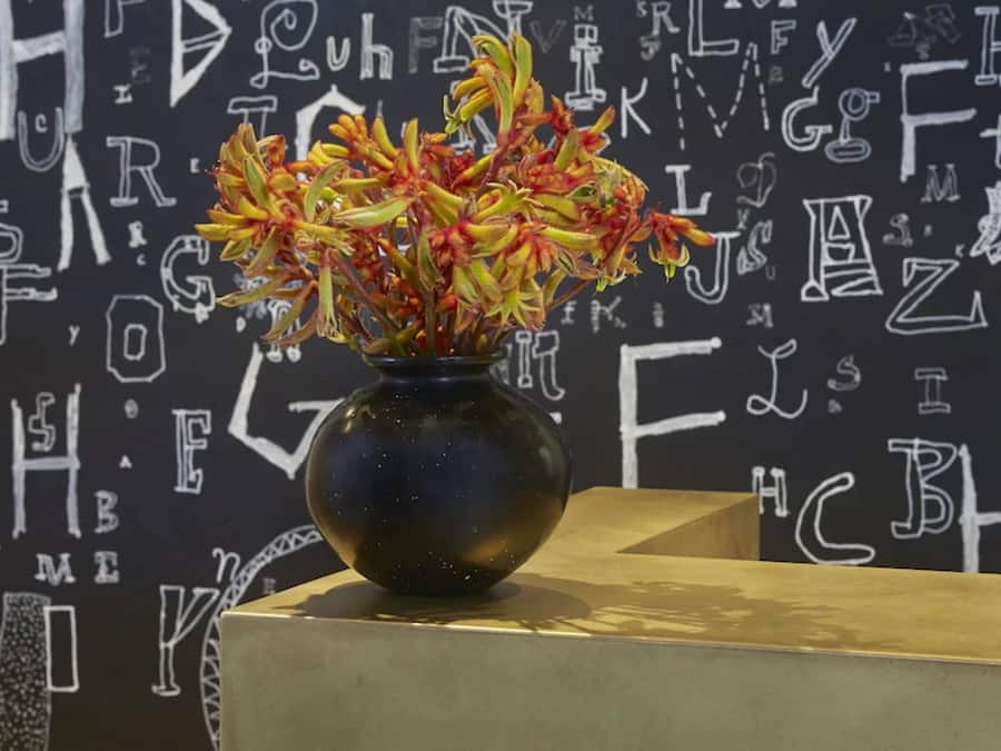 A vase with flowers on a table against a wall with white text.