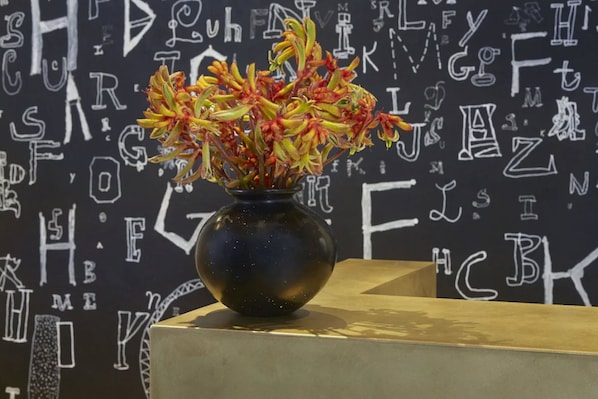 Un vase rempli de fleurs est posé sur une table, devant un mur sur lequel figure un texte blanc.