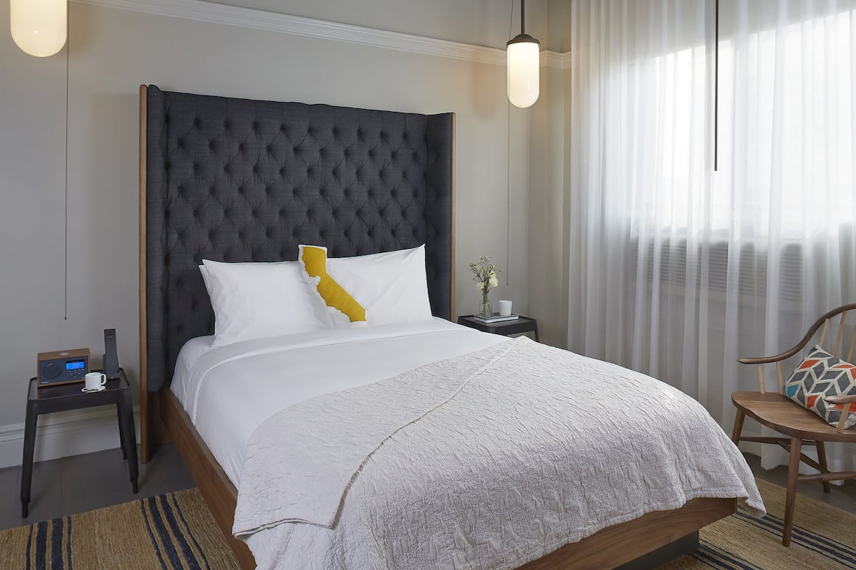 A modern hotel room with a large bed, a wooden headboard, a bedside table with a radio, a small table with a vase and a cup, and a chair with a patterned cushion.