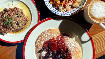 A table with three plates of food: a dish with a meat-based topping, a bowl of fruit salad, and a plate with pancakes topped with berries and powdered sugar.