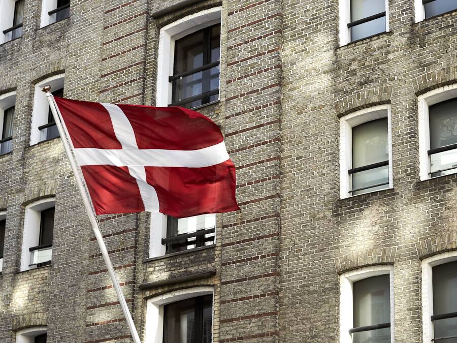 En flagstang med et flag foran en murstensbygning med mange vinduer.