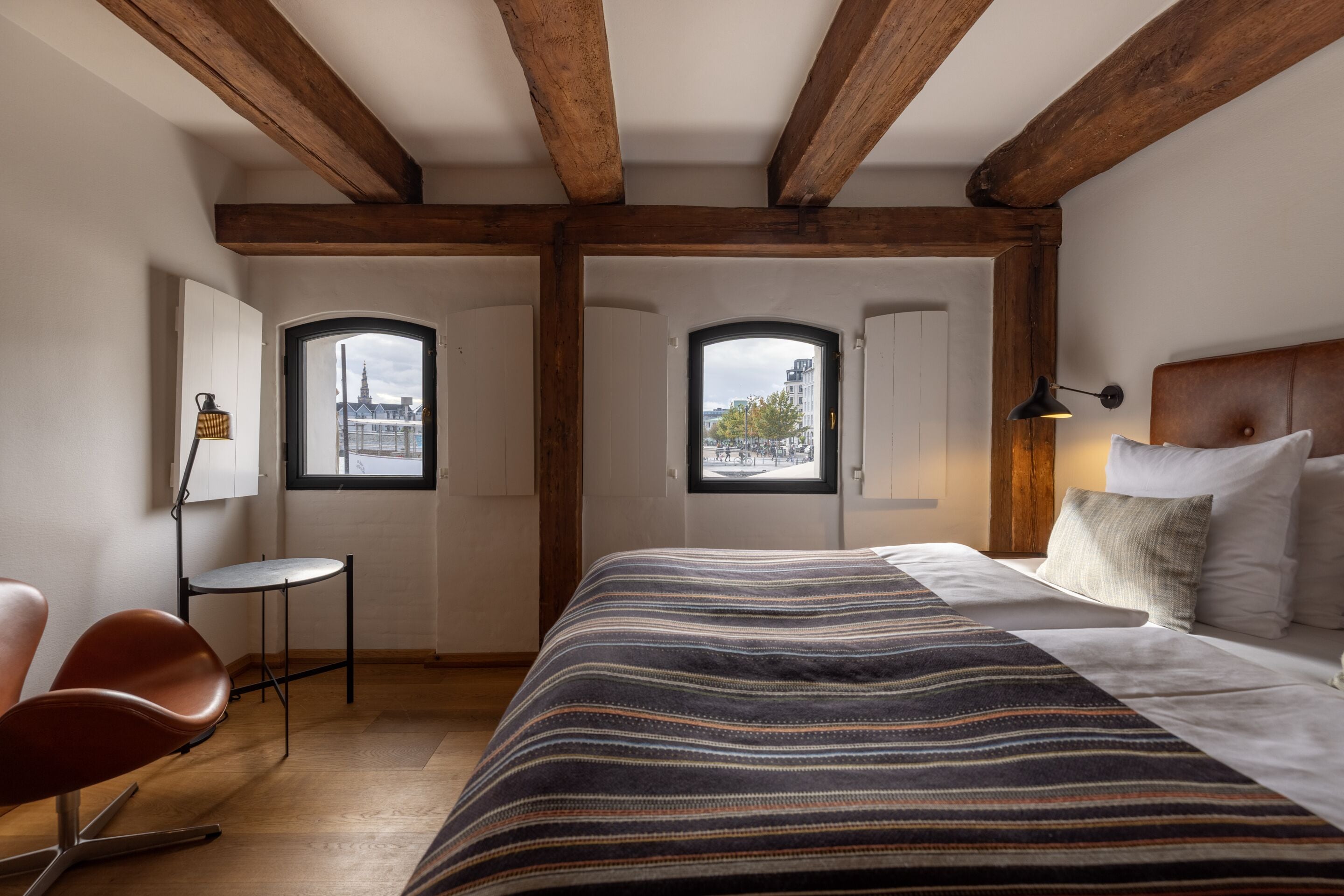 Un dormitorio con una cama grande, vigas de madera, dos ventanas con vistas a la ciudad, un escritorio y una silla.