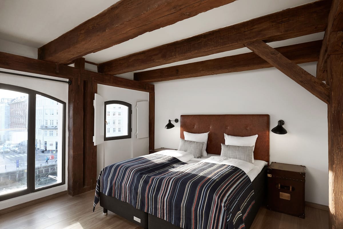 Une chambre avec un grand lit, des poutres apparentes, une vue sur la ville et une petite table de chevet.
