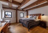 A bedroom with a bed, a desk, and a chair, wooden beams on the ceiling, and a window with a view of buildings.