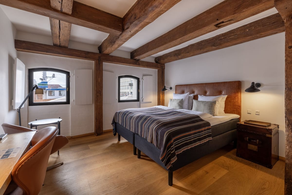 Une chambre avec un lit, un bureau et une chaise, des poutres en bois au plafond et une fenêtre donnant sur des immeubles.