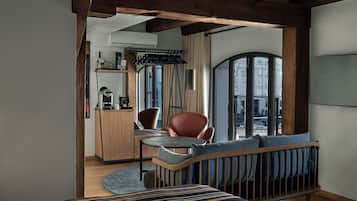 A modern hotel room with a wooden ceiling, a cozy seating area with a cushioned bench and a small table, a kitchenette, and a large window offering a city view.