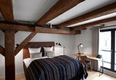 A bedroom with a large bed, wooden beams, a desk, and a window with curtains.