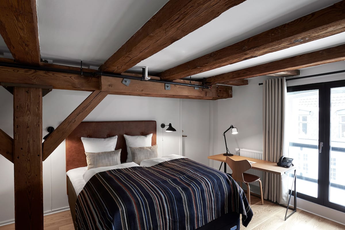 Une chambre avec un plafond en bois, un lit recouvert d’une couverture à rayures, un bureau avec une chaise et une fenêtre avec des rideaux.