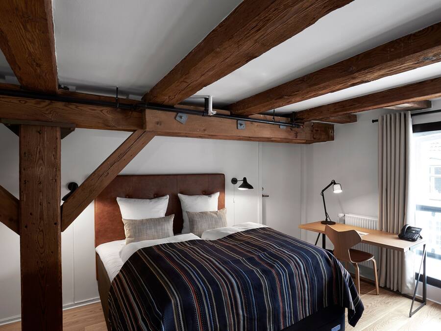 A bedroom with a large bed, wooden beams, a desk, and a window with curtains.